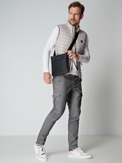 Ein Mann in einer hellen Pufferweste und einem weißen Langarmhemd hält eine schwarze Umhängetasche. Er trägt graue Cargohosen und weiße Turnschuhe. Er macht eine lässige Pose vor einem schlichten Hintergrund.