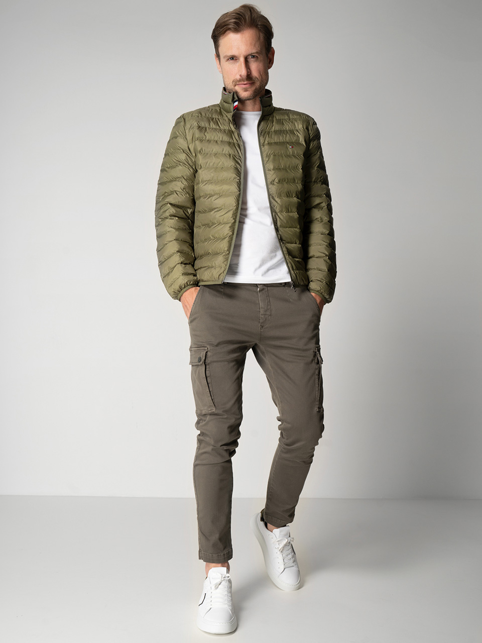 Eine Person in olivgrüner Pufferjacke, weißem T-Shirt, Khakihose und weißen Turnschuhen posiert vor einem schlichten weißen Hintergrund. Sie hat eine Hand in der Tasche und steht entspannt da.