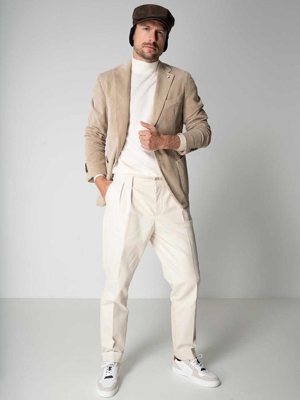 Eine Person in einem beige karierten Blazer, einem weißen Rollkragenpullover, cremefarbenen Bundfaltenhosen und weißen Sneakers steht selbstbewusst vor einem schlichten Hintergrund. Sie trägt außerdem eine braune Schiebermütze und blickt leicht zur Seite.