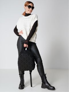 Eine stylische Frau mit schwarzer Sonnenbrille, einem weißen ärmellosen Pullover über einem schwarzen Rollkragenpullover und schwarzen Lederhosen posiert selbstbewusst. Sie hält einen schwarzen Rucksack und trägt schwarze Schnürstiefel. Sie steht vor einem schlichten Hintergrund.