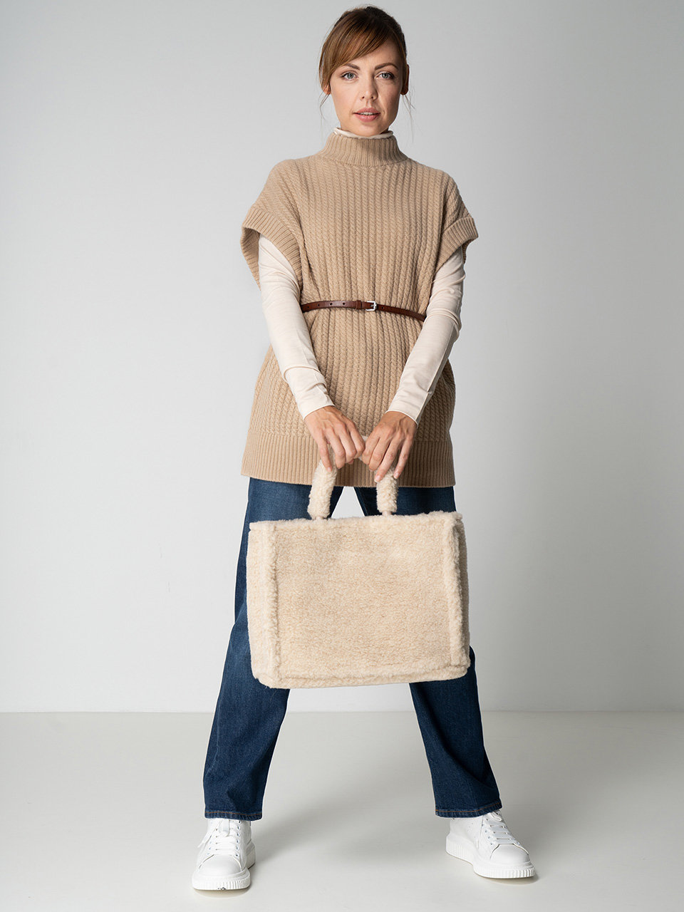 Eine Person in einem beigen Strickpullover, einem weißen Langarmshirt und blauen Jeans steht vor einem schlichten Hintergrund. Sie hält eine beigefarbene, flauschige Handtasche und trägt weiße Turnschuhe. Ein brauner Gürtel hält den Pullover in der Taille fest.