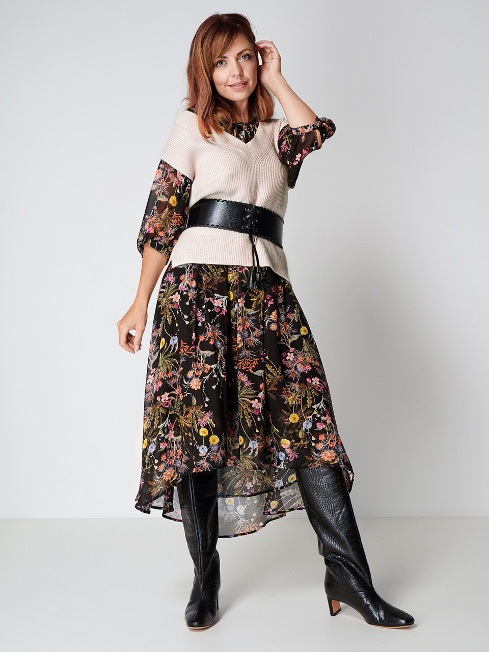 Eine Frau posiert selbstbewusst in einem geblümten Kleid, dazu eine beigefarbene Pullunderweste und einen breiten schwarzen Gürtel. Den Look rundet sie mit schwarzen kniehohen Stiefeln ab und steht vor einem schlichten weißen Hintergrund.