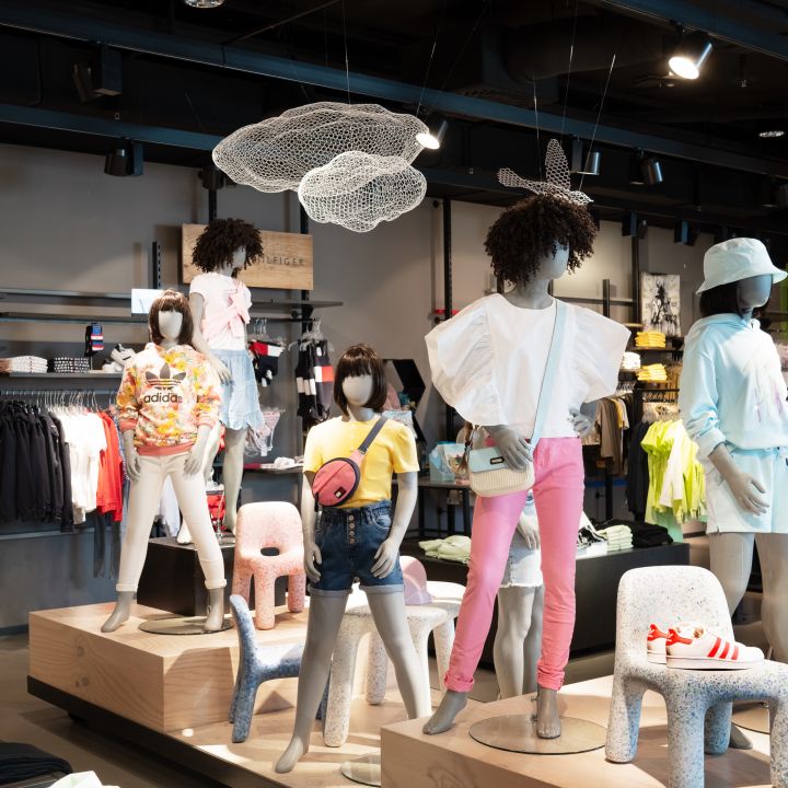 Ein Schaufenster in einem Kinderbekleidungsgeschäft mit Schaufensterpuppen in farbenfrohen Outfits, darunter rosa Hosen, ein weißes Kleid und Shorts mit einer rosa Tasche. Im Geschäft gibt es Kleiderständer und von der Decke hängen dekorative Drahtwolken.