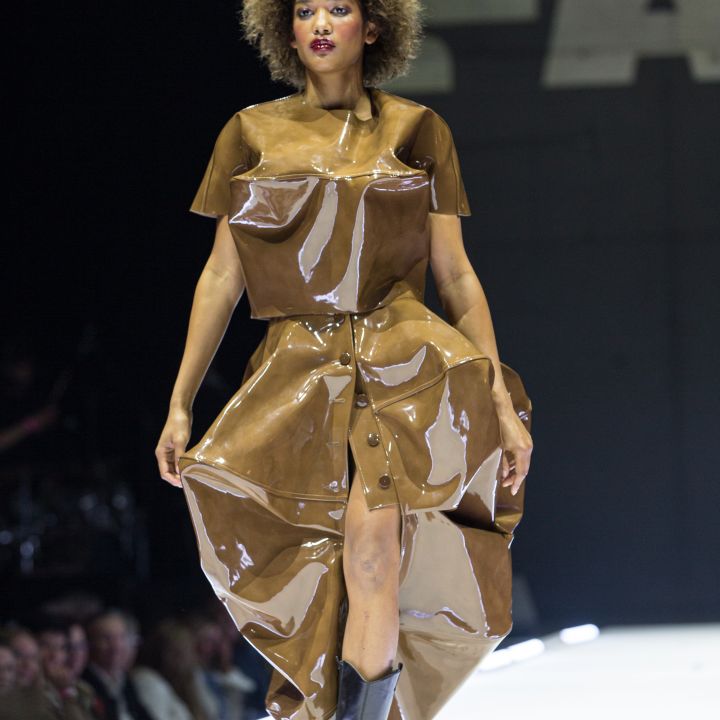 Ein Model läuft den Laufsteg entlang und trägt ein glänzendes, braunes, plastikartiges Oberteil und einen Rock, dazu schwarze Stiefel. Der Hintergrund ist dunkel, und man sieht teilweise ein Publikum, das die Modenschau verfolgt.