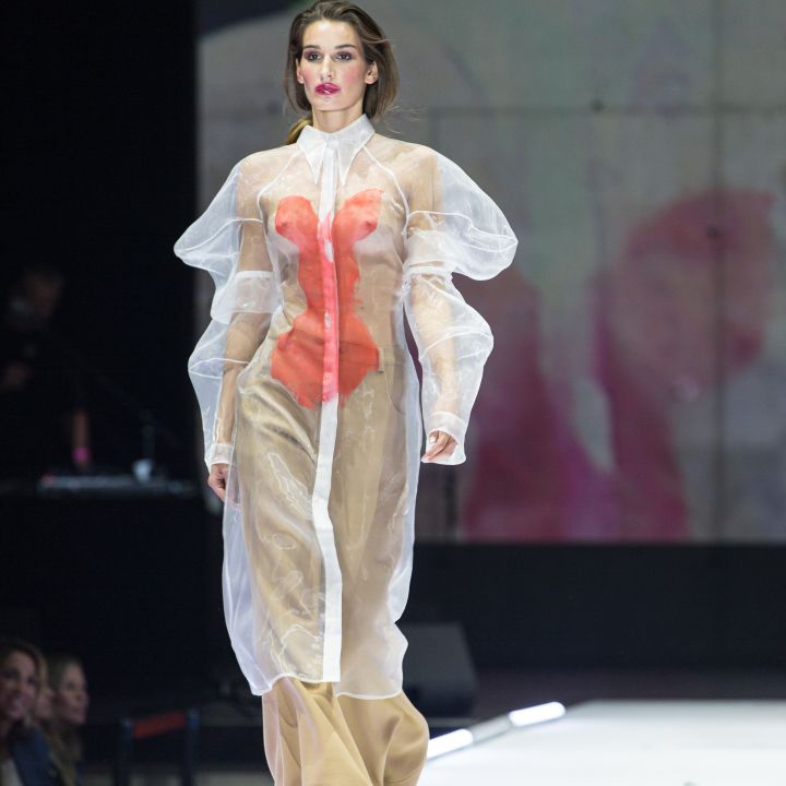 Ein Model läuft über einen Laufsteg und trägt ein transparentes, weißes Kleid mit Puffärmeln und orangefarbenem Innenteil sowie beigefarbene Hosen. Im Hintergrund ist ein verschwommenes Bild zu sehen, seitlich sind Zuschauer zu sehen.