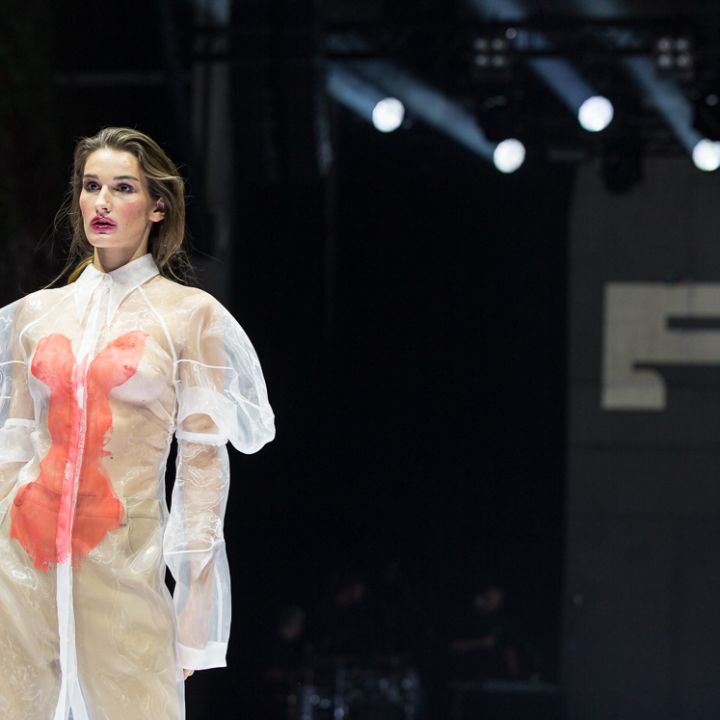 Ein Model läuft in einem transparenten, langärmeligen Kleid mit kräftigen roten Akzenten unter hellem Bühnenlicht über den Laufsteg. Der Hintergrund ist eine dunkle Kulisse mit einem großen, teilweise sichtbaren Schild.