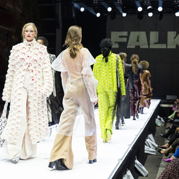 Models laufen in unterschiedlichen Modeoutfits, darunter einem Blumenensemble und einem transparenten Kleidungsstück, über einen hell erleuchteten Laufsteg. Ein großes Publikum schaut zu, im Hintergrund wird „FALK“ angezeigt.