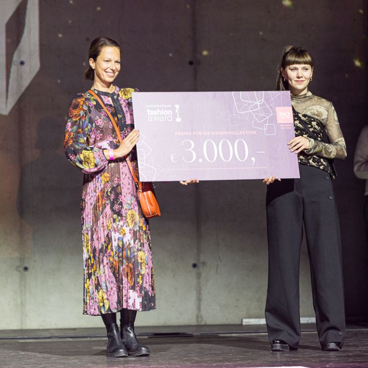 Zwei Frauen stehen bei einer Modepreisverleihung auf der Bühne und halten einen großen rosa Scheck über 3.000 Euro in der Hand. Eine trägt ein geblümtes Kleid, die andere eine schwarze Hose und ein Top. Im Hintergrund ist ein K&O-Logo zu sehen, eine weitere Person steht weiter hinten.