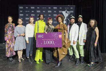 Eine Gruppe von zehn Personen steht auf einer Bühne, wobei eine Person einen großen lilafarbenen Preisscheck über 3.000 Dollar in der Hand hält. Sie tragen schicke, farbenfrohe Outfits. Hinter ihnen ist ein Hintergrund mit dem Namen der Veranstaltung zu sehen.
