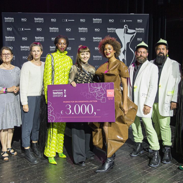 Eine Gruppe von zehn Personen steht auf einer Bühne, wobei eine Person einen großen lilafarbenen Preisscheck über 3.000 Dollar in der Hand hält. Sie tragen schicke, farbenfrohe Outfits. Hinter ihnen ist ein Hintergrund mit dem Namen der Veranstaltung zu sehen.