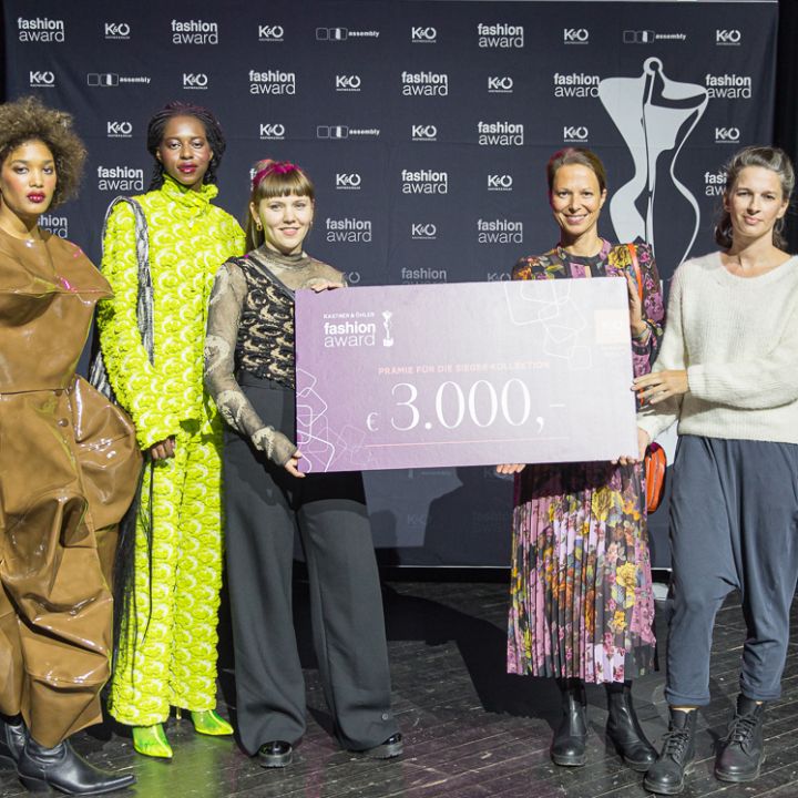Fünf Personen stehen auf einer Bühne und halten einen großen Scheck über 3.000 Euro in den Händen. Im Hintergrund ist das Logo eines Fashion Awards zu sehen. Sie sind in stylische, farbenfrohe Outfits gekleidet und scheinen zu feiern.