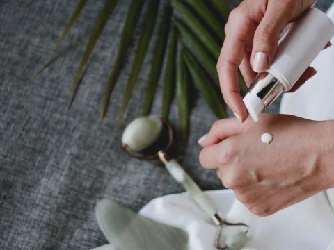Eine Person trägt eine kleine Menge Creme auf ihren Handrücken auf. In der Nähe befinden sich ein Jaderoller und ein grünes Blatt auf einem grauen Stoffhintergrund.