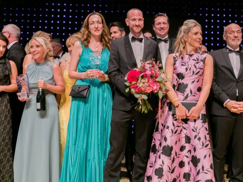 Eine Gruppe von Menschen in formeller Kleidung steht bei einer Veranstaltung eng beieinander. Einige halten Blumen oder Gläser in den Händen und alle lächeln. Im Hintergrund ist eine Wand mit kleinen, gleichmäßig verteilten Lichtern zu sehen.