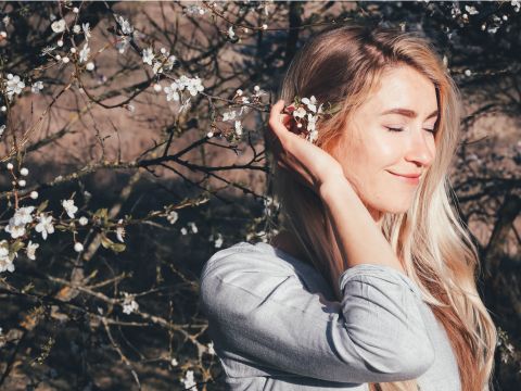 Eine Frau mit langen blonden Haaren steht mit geschlossenen Augen im Freien und lächelt sanft. Sie hält einen Zweig mit weißen Blüten an ihr Ohr. Das Sonnenlicht wirft einen warmen Schein und verschwommene Zweige und Blüten füllen den Hintergrund.