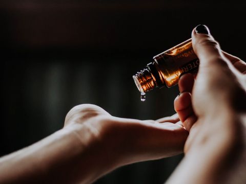 Eine Person gießt ätherisches Öl aus einer kleinen Bernsteinflasche in ihre offene Handfläche. Der Hintergrund ist leicht verschwommen, wodurch Hand und Flasche hervorgehoben werden. Die Szene suggeriert einen Moment der Selbstpflege oder Aromatherapie.