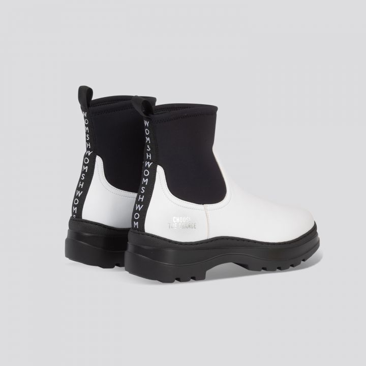 Ein Paar stylische Stiefeletten mit weißer Lederfront und schwarzer Gummisohle hinten, mit Laschen mit der Aufschrift „WOMSH“ und einem weißen Text auf der Ferse mit der Aufschrift „CHOOSE THE CHANGE“. Die Stiefel haben dicke schwarze Sohlen.