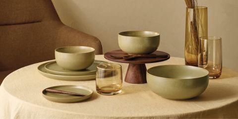 Eine beige Tischdecke umgibt ein minimalistisches Esstischset mit grünen Schüsseln, Tellern und Holzbesteck. Ein Holzständer hält eine Schüssel. Bernsteinfarbene Gläser und eine hohe Vase mit getrockneten Pflanzen sorgen für Wärme. Im Hintergrund steht ein brauner Stuhl.