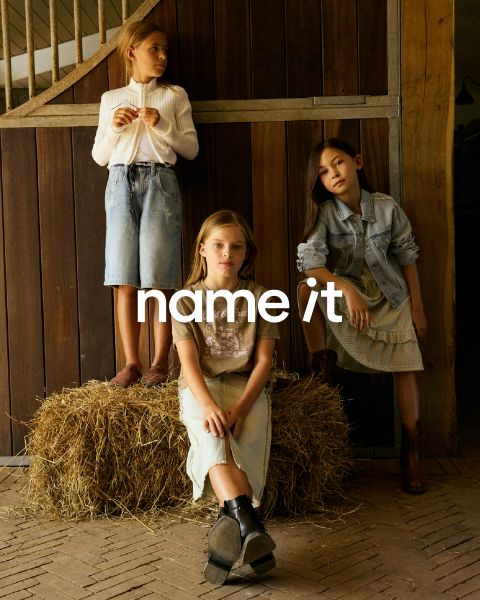 Drei Kinder in legerer Kleidung posieren in einer rustikalen Umgebung mit Holzwänden und Heuballen. Zwei stehen, während eines sitzt. Es trägt verschiedene Jeans und neutrale Farben. In der Mitte ist der Text „Name it“ eingeblendet.