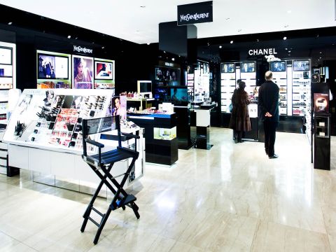 Ein heller, moderner Kosmetikladen mit beleuchteten Auslagen für Marken wie Yves Saint Laurent und Chanel. Zwei Personen stöbern in den Produkten, im Vordergrund steht ein Schminkstuhl mit dem Yves Saint Laurent-Logo.