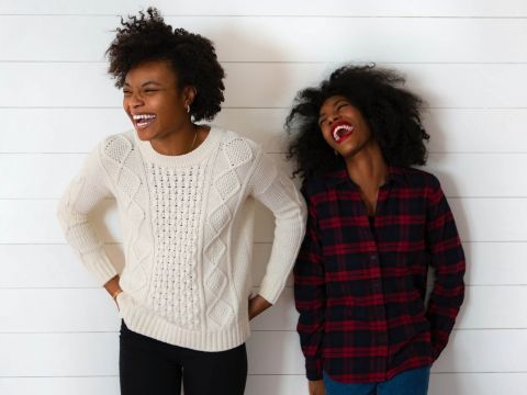 Zwei Menschen stehen vor einer weißen Wand mit Überlappungsbrettern und lachen fröhlich. Einer trägt einen cremefarbenen Pullover, der andere ein rot-schwarz kariertes Hemd. Ihre Ausdrücke vermitteln Freude und Kameradschaft.