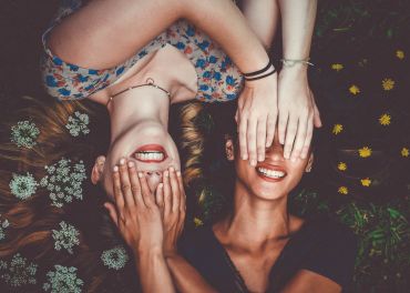 Zwei Frauen liegen mit Blumen im Gras und lächeln breit. Sie bedecken einander mit den Händen die Augen. Eine trägt ein geblümtes Kleid, die andere ein dunkles Oberteil. Auf dem Gras liegen verstreut gelbe Blumen.