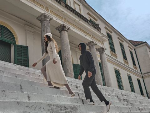 Zwei Menschen gehen Steintreppen vor einem historischen Gebäude mit grünen Fensterläden und großen Säulen hinauf. Einer trägt ein weißes Kleid und einen Hut, der andere einen dunklen Kapuzenpullover und Hosen. Der Himmel ist teilweise bewölkt.