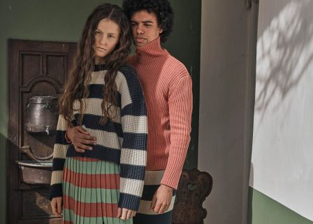 Eine Frau mit langen Haaren trägt einen gestreiften Pullover und Rock und steht vor einem Mann in einem rosa Rollkragenpullover. Sie posieren drinnen neben einer rustikalen Holztür, wobei sanftes Tageslicht Schatten an die Wand wirft.