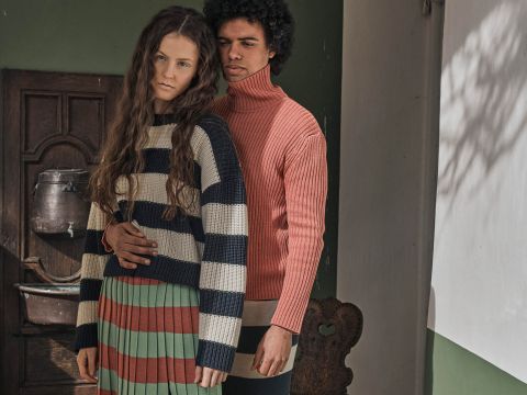 Eine Frau mit langen Haaren trägt einen gestreiften Pullover und Rock und steht vor einem Mann in einem rosa Rollkragenpullover. Sie posieren drinnen neben einer rustikalen Holztür, wobei sanftes Tageslicht Schatten an die Wand wirft.