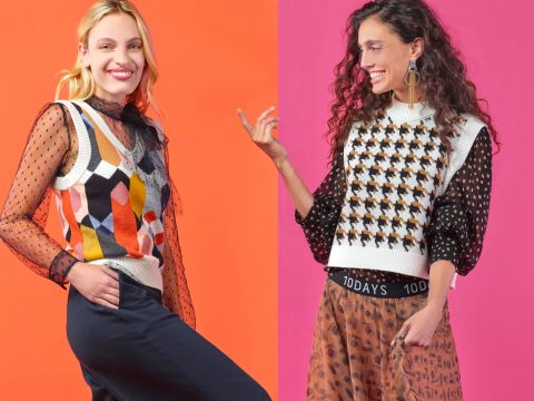 Zwei Frauen stehen lächelnd vor einem lebhaften, gespaltenen Hintergrund, wobei eine Seite orange und die andere rosa ist. Sie tragen farbenfrohe, gemusterte Oberteile; eine trägt eine Weste mit geometrischem Muster, die andere ein Hahnentrittmuster.