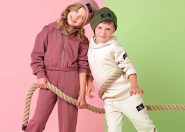 Zwei Kinder lächeln und halten ein Seil vor einem gespaltenen rosa und grünen Hintergrund. Das Mädchen trägt einen rosa Trainingsanzug und eine passende Mütze, während der Junge einen weißen Trainingsanzug und eine grüne Mütze trägt. Beide Kinder haben Markenaufnäher auf ihren Outfits.