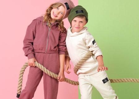 Zwei Kinder lächeln und halten ein Seil vor einem gespaltenen rosa und grünen Hintergrund. Das Mädchen trägt einen rosa Trainingsanzug und eine passende Mütze, während der Junge einen weißen Trainingsanzug und eine grüne Mütze trägt. Beide Kinder haben Markenaufnäher auf ihren Outfits.