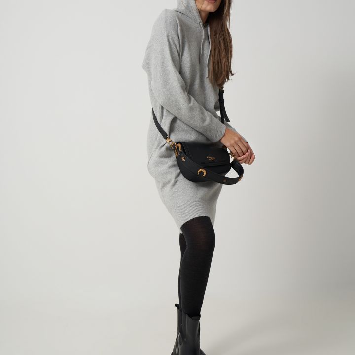 Eine Person in einem grauen Kapuzenkleid und schwarzen Kniestrümpfen posiert vor einem schlichten Hintergrund. Sie trägt schwarze Stiefeletten und eine schwarze Umhängetasche, hat lange Haare und einen entspannten Gesichtsausdruck.