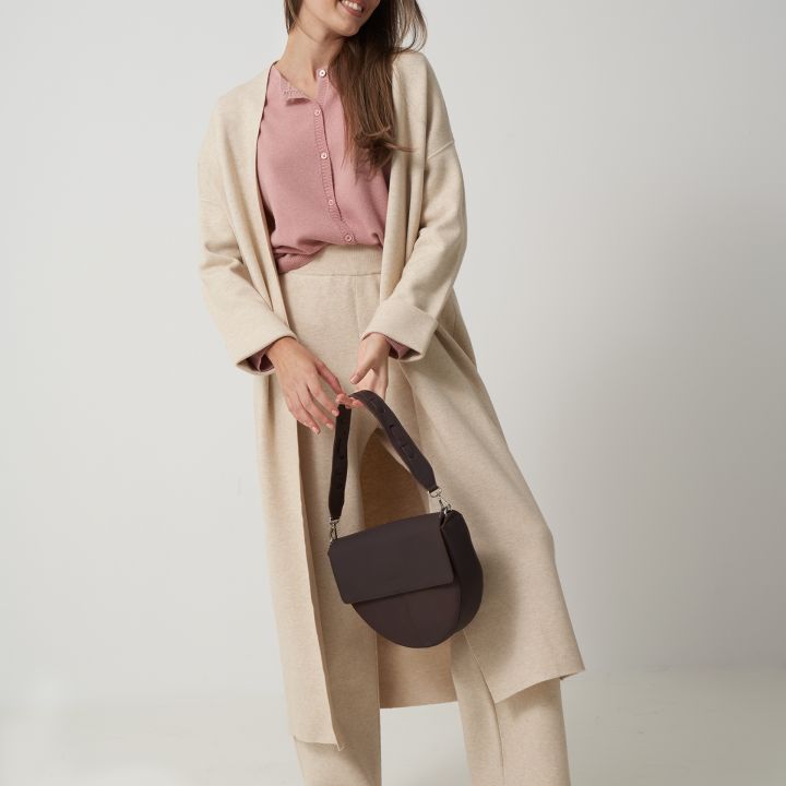 Eine Frau lächelt und trägt einen langen beigen Mantel über einer rosa Bluse und beigen Hosen. Sie hält eine dunkelbraune Handtasche in der Hand und trägt weiße Schuhe. Der Hintergrund ist schlicht hellgrau.