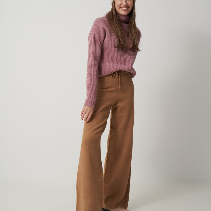 Eine Person in einem rosa Pullover, beigen Hosen mit weitem Bein und einer Strickmütze steht lächelnd vor einem schlichten weißen Hintergrund. Ihr Outfit wird durch hellbraune Stiefel ergänzt.