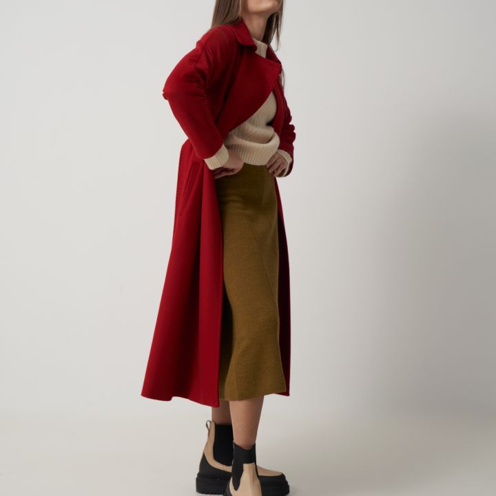 Eine Frau steht selbstbewusst vor einem schlichten Hintergrund. Sie trägt einen langen roten Mantel über einem beigen Pullover und einem olivgrünen Rock. Ihr Outfit wird durch beige und schwarze Plateaustiefel vervollständigt. Ihre Hände liegen auf ihren Hüften.