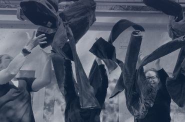 Zwei Menschen werfen vor einem Betonhintergrund freudig Jeans in die Luft. Beide tragen legere schwarze Kleidung und scheinen zu feiern oder zu spielen. Über ihnen sind Jeans im Flug zu sehen. Die Szene ist lebhaft und dynamisch.