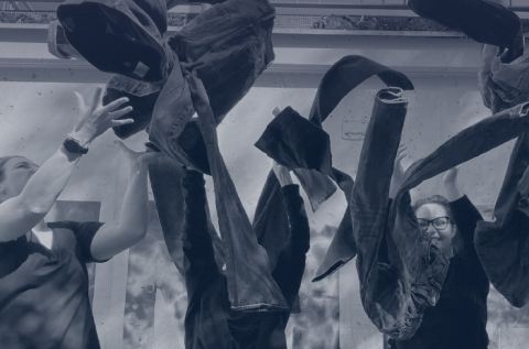 Zwei Menschen werfen vor einem Betonhintergrund freudig Jeans in die Luft. Beide tragen legere schwarze Kleidung und scheinen zu feiern oder zu spielen. Über ihnen sind Jeans im Flug zu sehen. Die Szene ist lebhaft und dynamisch.