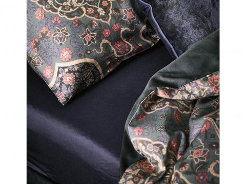 Eine Nahaufnahme eines Bettes mit dunklen, reich gemusterten Bettlaken und Kissenbezügen mit Blumenmuster in Rot-, Gold- und Grüntönen. Die Bettwäsche umfasst ein strukturiertes Samtkissen und eine passende Decke, die eine luxuriöse und gemütliche Atmosphäre schaffen.
