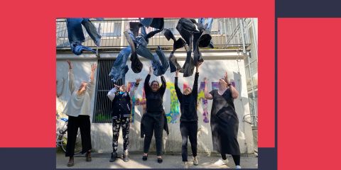 Fünf Menschen stehen vor einem Gebäude und werfen fröhlich Jeans in die Luft. Sie sind draußen, mit Graffiti an der Wand hinter ihnen, und genießen einen sonnigen Tag.
