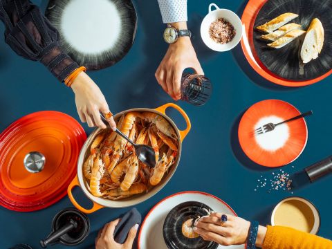 Eine Draufsicht auf einen Esstisch mit Leuten, die servieren und essen. In der Mitte steht ein Topf mit Meeresfrüchten, umgeben von roten und schwarzen Tellern, Brot, einer Schale mit Salz und Getränken. Hände greifen nach Essen und Getränken und schaffen so eine lebhafte Essszene.