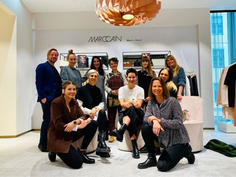 Eine Gruppe von zehn Personen in Freizeit- und Businesskleidung posiert und lächelt in einem schicken Einzelhandelsgeschäft. Hinter ihnen befindet sich eine Ausstellungswand mit Regalen und Kleidungsstücken. Über ihnen hängt eine moderne, spiralförmige Leuchte.