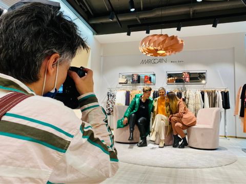 Ein Fotograf macht ein Foto von drei Personen, die in einem Modegeschäft auf einem Sofa sitzen. Das Geschäft heißt Marc Cain und hat im Hintergrund Kleiderständer und eine stilvolle Lampe darüber. Die Personen lächeln, einer trägt eine grüne Jacke.