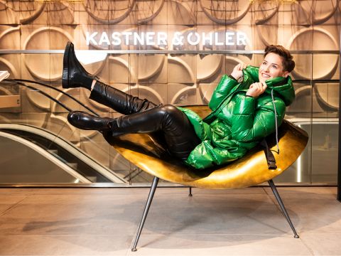 Eine Person in einer glänzenden grünen Jacke und schwarzen Lederhosen posiert verspielt auf einem stilvollen, modernen goldenen Stuhl. Sie hat die Beine hochgelegt und lächelt, im Hintergrund ist „Kastner & Öhler“ zu sehen.