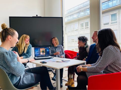 Eine Gruppe von sechs Personen sitzt in einem modernen Büro um einen Tisch und diskutiert. In der Mitte steht ein geöffneter Laptop, und große Fenster sorgen für Tageslicht. Alle scheinen zu lächeln und entspannt zu sein.