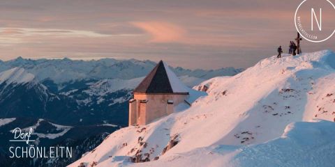 Schneebedeckte Berglandschaft bei Sonnenuntergang mit einer kleinen Hütte neben einer Gruppe von Menschen auf einem Bergrücken. Rosa und orange Farbtöne erhellen den Himmel und heben die schneebedeckten Gipfel hervor. Textüberlagerung lautet „Dorf SCHÖNLEITN“. Logo in der Ecke.