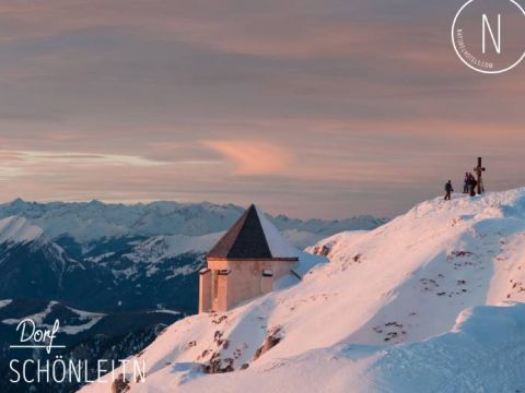 Ein schneebedeckter Berggipfel bei Sonnenaufgang, auf dem sich ein kleines Gebäude mit Spitzdach befindet. Zwei Menschen stehen neben einem Kreuz und blicken auf eine weite Bergkette in der Ferne unter einem rosa und orangefarbenen Himmel.