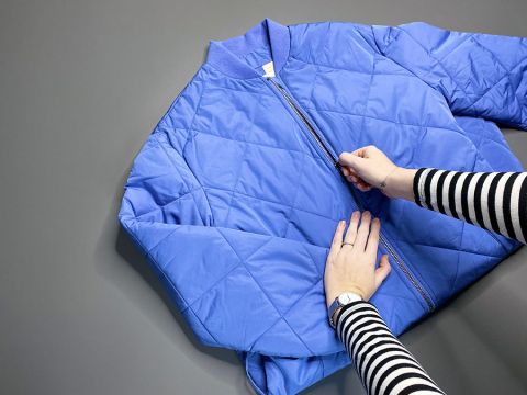 Eine Person in einem gestreiften Langarmhemd zieht den Reißverschluss einer blauen Steppjacke zu, die flach auf einer grauen Oberfläche liegt.