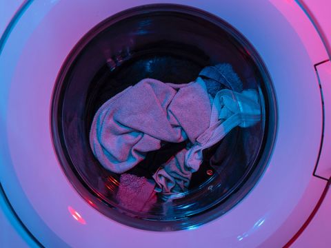 Eine Nahaufnahme einer Waschmaschinentrommel mit verschiedenen Kleidungsstücken, darunter ein hellgraues Handtuch und dunkle Kleidungsstücke. Die Szene wird mit rosa und blauem Licht beleuchtet, wodurch ein farbenfroher Effekt entsteht.