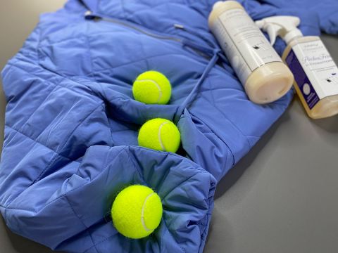 Ausgebreitet liegt eine blaue Steppjacke, in deren Falten drei gelbe Tennisbälle teilweise eingesteckt sind. Daneben stehen auf einer grauen Fläche zwei Flaschen mit Reinigungsmitteln.