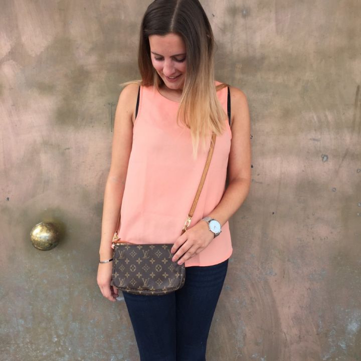 Eine Frau mit langen Haaren steht lächelnd da und schaut nach unten. Sie trägt ein rosa ärmelloses Top und dunkle Jeans. Sie trägt eine kleine Designertasche und trägt eine Uhr und ein Armband. Der Hintergrund ist eine strukturierte Wand in subtilen Erdtönen.